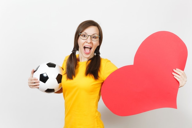 Smiling European woman, fun pony tails, football fan or player in glasses, yellow uniform hold classic soccer ball, red heart isolated on white background. Sport, football, healthy lifestyle concept.