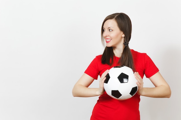 Smiling european young woman, two fun pony tails, football fan\
or player in red uniform hold classic soccer ball isolated on white\
background. sport play football, healthy lifestyle concept. side\
view