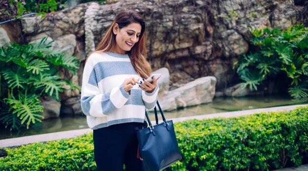 Smiling excited adult female in denim clothes with bag chilling in botanical garden with vegetation