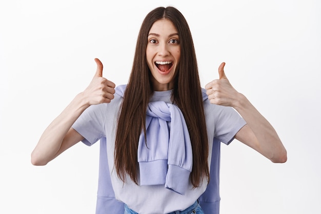 Smiling excited girl shows thumbs up in approval, agree and like good product, prause amazing job, excellent work or perfect choice, standing over white wall