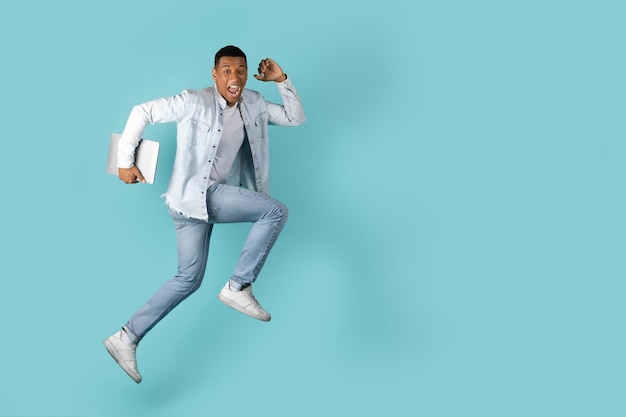 Smiling excited millennial african american guy with open mouth with laptop jumping in air to empty space