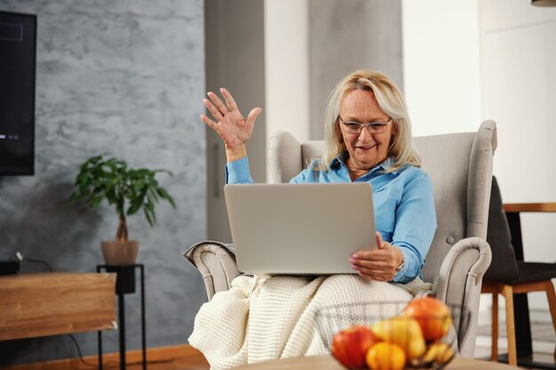 Sorridente, eccitata donna senior seduta in poltrona a casa e guardando il computer portatile