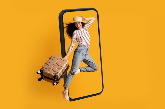 Smiling excited young arab lady tourist in hat with suitcase jumping near huge smartphone ready for travel