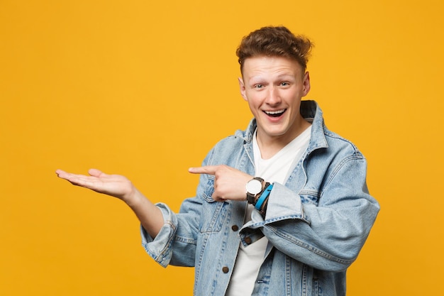 Smiling excited young man in denim casual clothes pointing hand, index finger aside isolated on yellow orange wall background in studio. people sincere emotions, lifestyle concept. mock up copy space