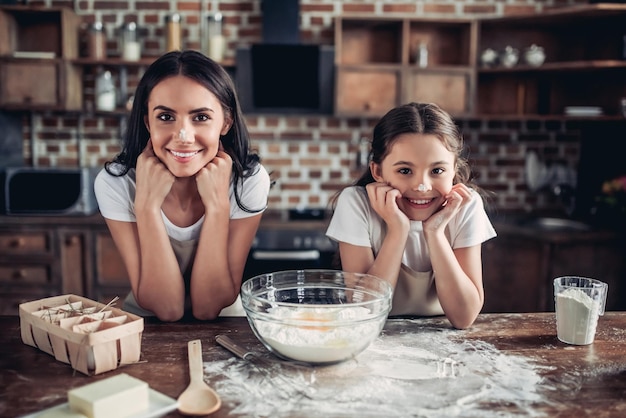 Smiling family of daughter and mother with noses in flour looking at the camera on the kitchen