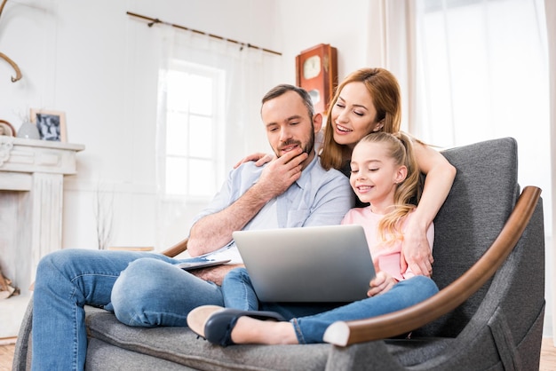 Улыбающаяся семья с одним ребенком сидит на диване и пользуется ноутбуком