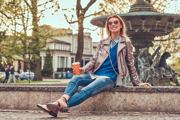 웃고 있는 세련된 금발 여성은 야외에서 휴식을 취하고 도시 공원의 벤치에 앉아 테이크아웃 커피를 마십니다.