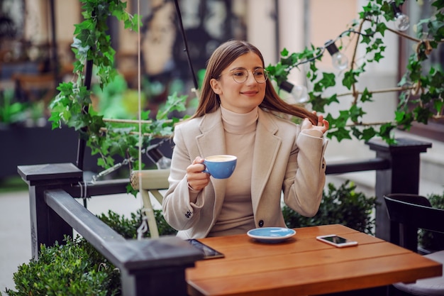 Улыбающаяся модная девушка колледжа сидит в кафе на открытом воздухе, держит чашку кофе и делает перерыв между лекциями.