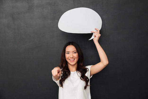 Smiling female asian holding blank speech bubble above her head and pointing finger on camera, isolated over dark gray wall