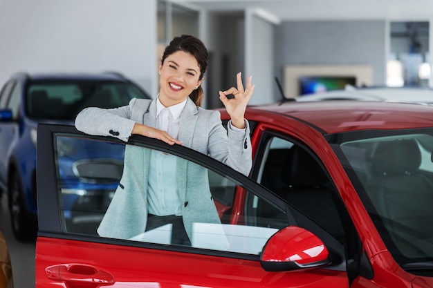 Smiling female car seller leaning on car while standing in car salon and shoving okay gesture with hand