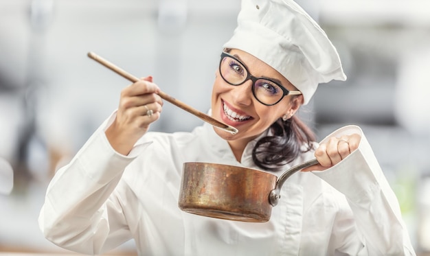 La chef femminile sorridente in abito professionale assaggia il cibo dal cucchiaio di legno che tiene una pentola di rame