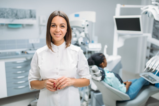 Smiling female dentist, dental clinic, patient in chair on background. Woman in dentistry cabinet, stomatology, teeth care