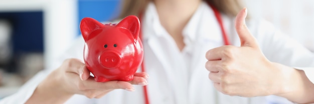 Smiling female doctor holding red pig piggy bank and thumbs up