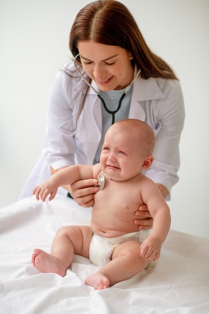 Smiling female doctor supporting a scared baby seated on the couch during the chest auscultation