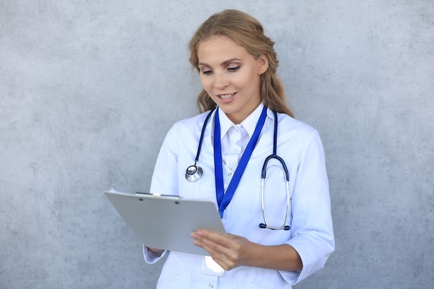 회색 배경 위에 격리된 건강 카드를 들고 청진기를 들고 웃고 있는 여성 의사.