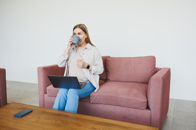 Smiling female freelancer in casual clothes sitting in comfortable sofa in modern cafeteria at table with laptop and phone talking on mobile phone remote work