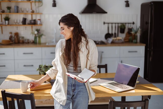 Foto una donna freelance sorridente con un auricolare che tiene un quaderno e una tazza vicino al portatile in cucina a casa