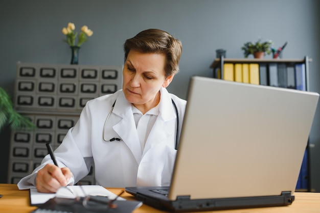 Улыбающаяся женщина-медицинский работник делает документы и использует ноутбук во время работы в кабинете врача