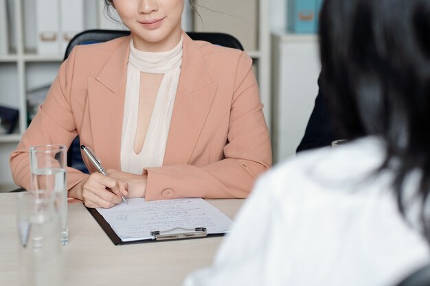 사진 면접에서 지원자와 이야기할 때 문서에 메모를 하는 웃는 여성 인사 관리자