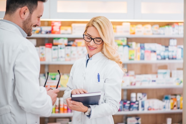 Smiling female pharmacist beggining to work at modern pharmacy, colleague giving her prescription book.