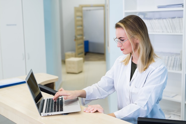 Smiling female physician using computer in her office in hospital. Young attractive Caucasian woman doctor therapist wearing medical uniform sitting at the desk