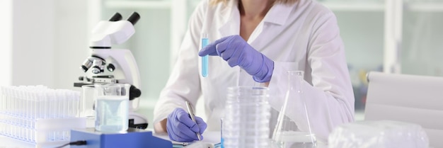 Smiling female scientist holding test tube with blue liquid and making notes chemist conducts