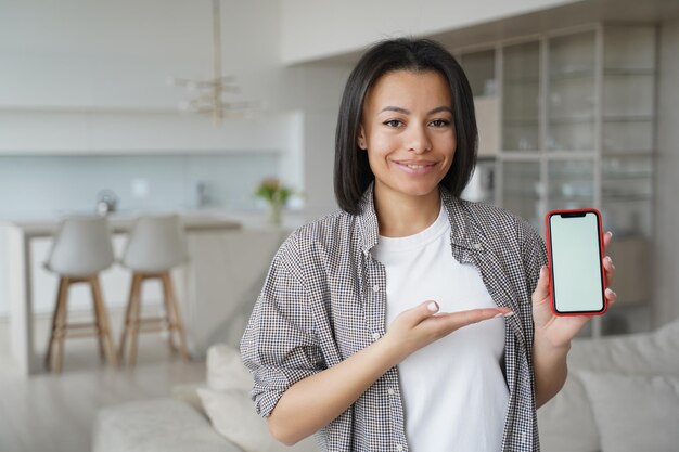 Smiling female show phone with blank mockup screen advertise\
mobile apps at home online store offer