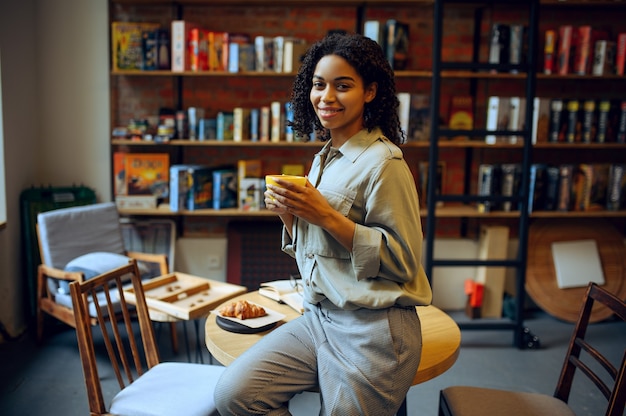 Smiling female student in library cafe. Woman with coffee and croissants poses at the table , education and knowledge. Girl studying in campus