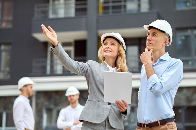 Smiling female supervisor with a ceramic tile in her hand pointing at something to her focused colleague in headwear