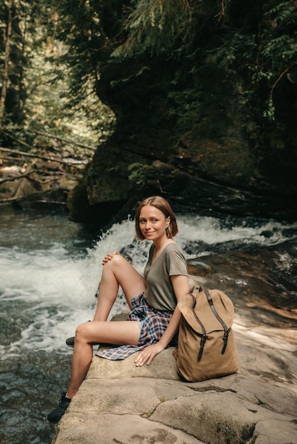 Photo smiling female traveler with backpack resting while sitting by the mountain river