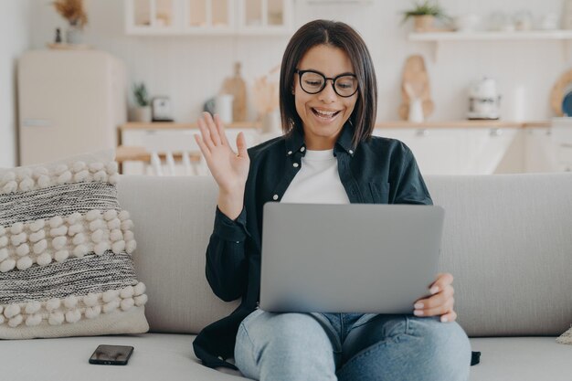 smiling female waving hand to laptop greeting teacher at lesson sitting on couch at home elearning
