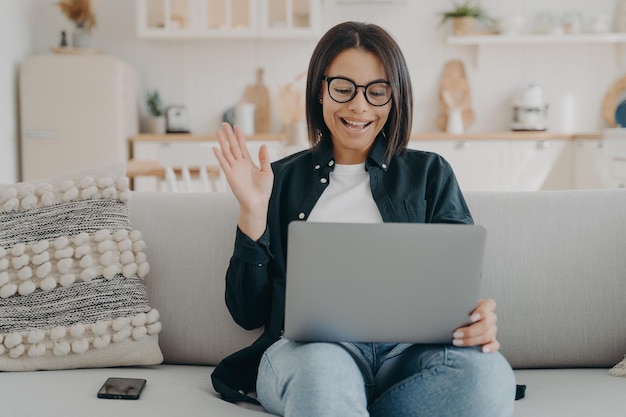 Smiling female waving hand to laptop greeting teacher at lesson sitting on couch at home Elearning