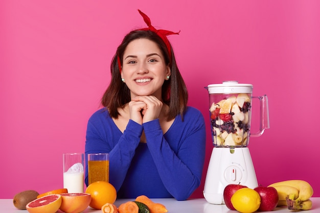 Smiling female, wears red headband, blue jumper, holds her head with arms, uses different ingredients, fruit and vegetables for preparing fresh smothie, leads healthy lifestyle. Dieting concept.