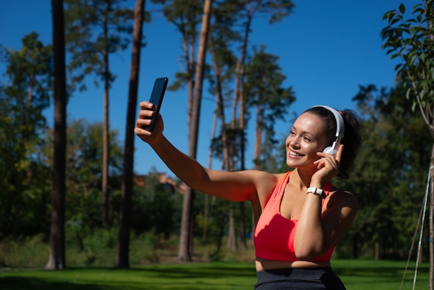 Smiling fit woman with headphones makes selfie while having reat in the beautiful park. Relaxation outdoor.