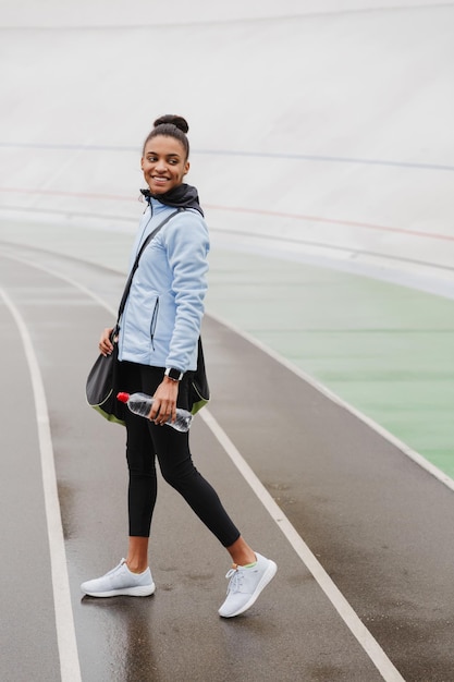 Smiling fit young african sportswoman carrying sportsbag standing at the stadium, holding water bottle