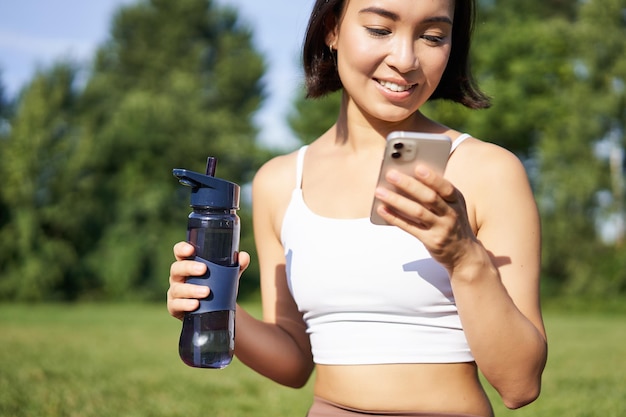 Smiling fitness girl drinks water checks her app on smartphone\
and looks happy stays hydrated on fre