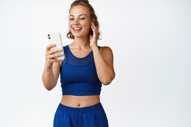 Smiling fitness woman listening music in wireless headphones. Sportswoman listening to sports instructor on smartphone app, workout in earphones on white.