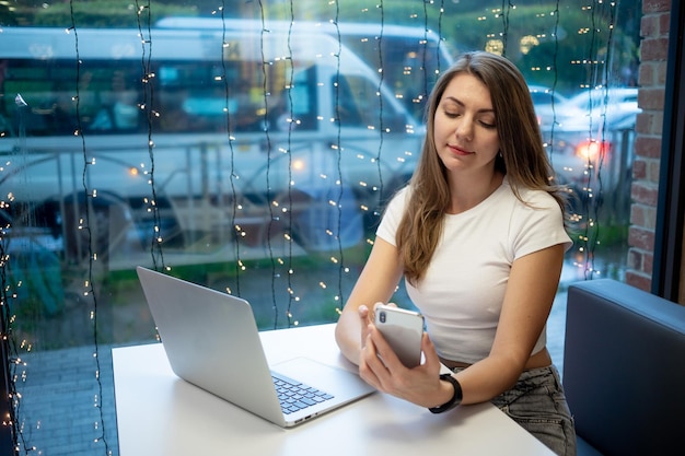 Sorridente ragazza freelance che lavora al computer portatile in un bar e fa videochiamate o parla al telefono la giornata lavorativa di una giovane donna o studente libera