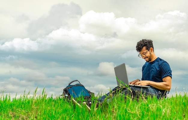 Uomo sorridente freelance seduto con laptop e zaino in campo ritratto di un uomo seduto sull'erba del campo che lavora con il suo laptop concetto di uomo freelance che lavora dal campo