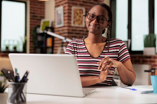 Smiling freelancer sitting at workplace desk, working on\
laptop, looking at camera. successful remote employee at home\
office table, online courses young cheerful student portrait