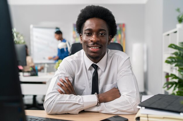 Smiling friendly afro man sits on desk wearing white shirt and tie in background scenery of company