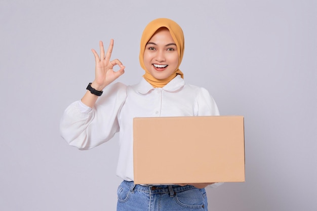 Smiling friendly Asian Muslim Woman in white shirt holding package box and showing okay sign isolated on white background Shopping and delivery concept