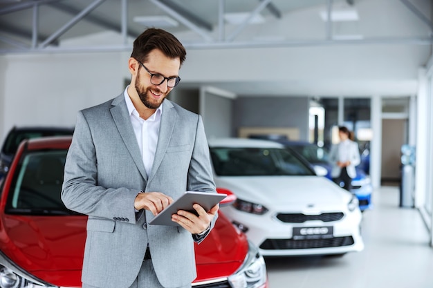 Smiling, friendly car seller standing in car salon and using tablet to check on new messages customers post on internet