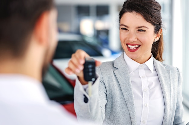 Photo smiling, friendly car seller standing in car salon with a customer and handing him car keys of best selling car.