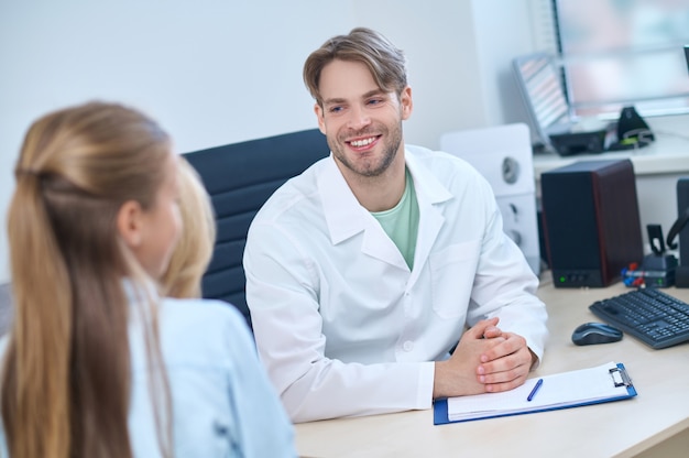 Smiling friendly handsome blond Caucasian male otorhinolaryngologist receiving a young female patient in his office