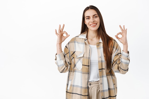 Smiling friendly woman shows okay sign, praising good quality, recommending company or promo deal, like something, give positive feedback, standing over white wall