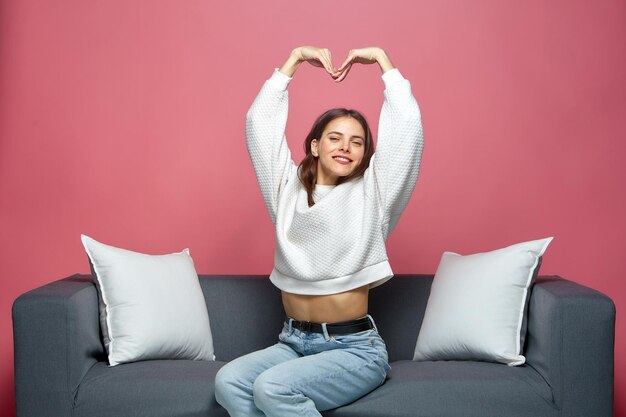 Smiling friendly young girl stretching hands up showing heart feeling love sitting on sofa women