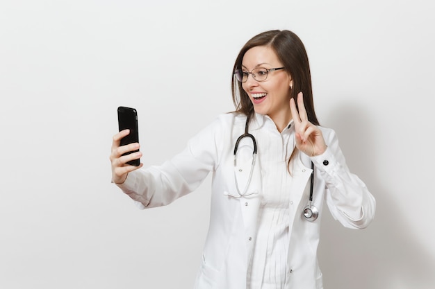 Smiling fun beautiful young doctor woman with stethoscope, glasses isolated on white background. Female doctor in medical gown doing selfie on mobile phone. Healthcare personnel, health, medicine conc