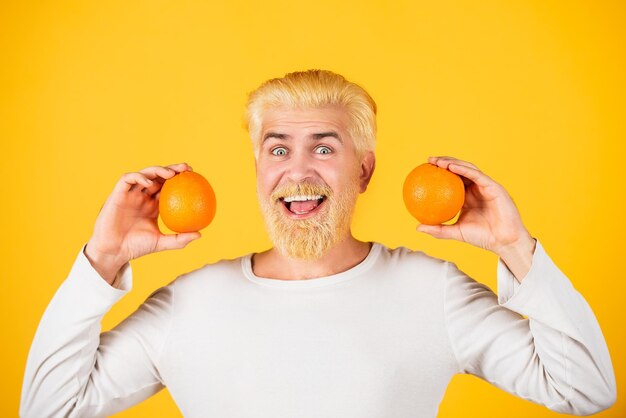 Улыбающийся забавный мужчина с апельсинами в руках Красивый молодой человек держит свежий натуральный апельсин Человек, наслаждающийся фруктовым освежающим апельсином на оранжевом фоне изолирован