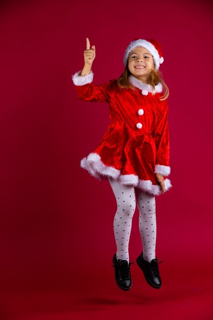 Smiling funny schoolgirl in red Christmas costume with red Santa's hat jumping on red colored wall with plenty of space. Kid points up the finger. . 